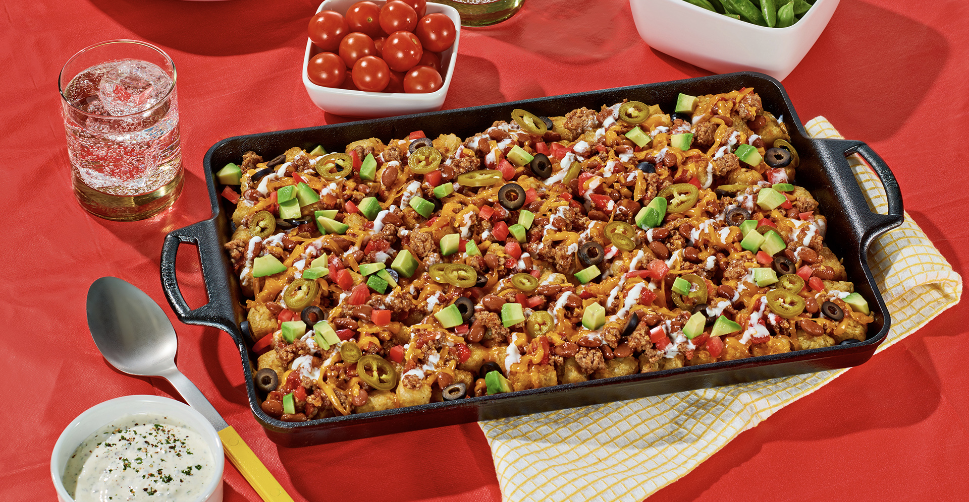 Chili Cheese totchos on a black tray sitting on a red table next to containers of cherry tomatoes and snap peas and a drink.