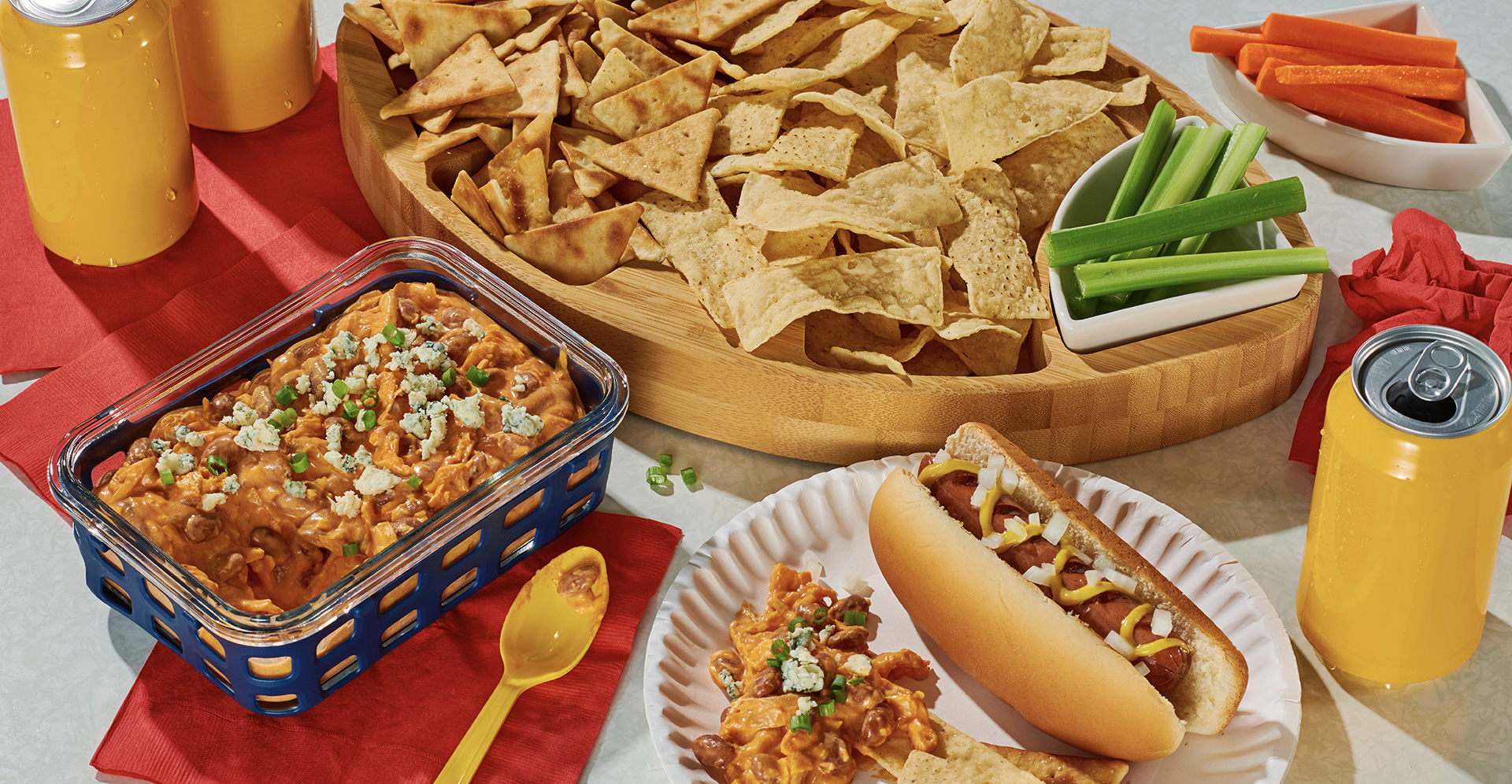 A tray of tortilla chips next to a dish of Buffalo chicken chili bean dip and a plate with a hotdog on it
