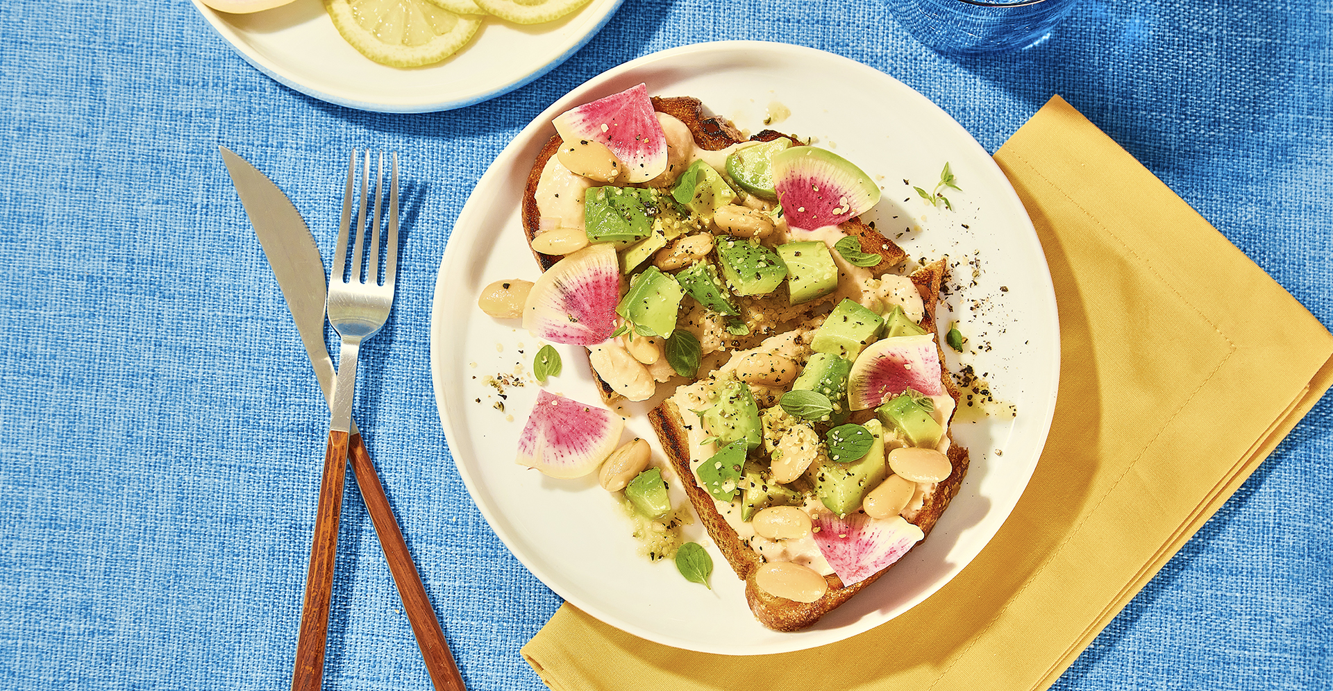 A large piece of toast with cannellini cream, avocado/bean mix, and shaved radish on a white plate, sitting on a blue table.