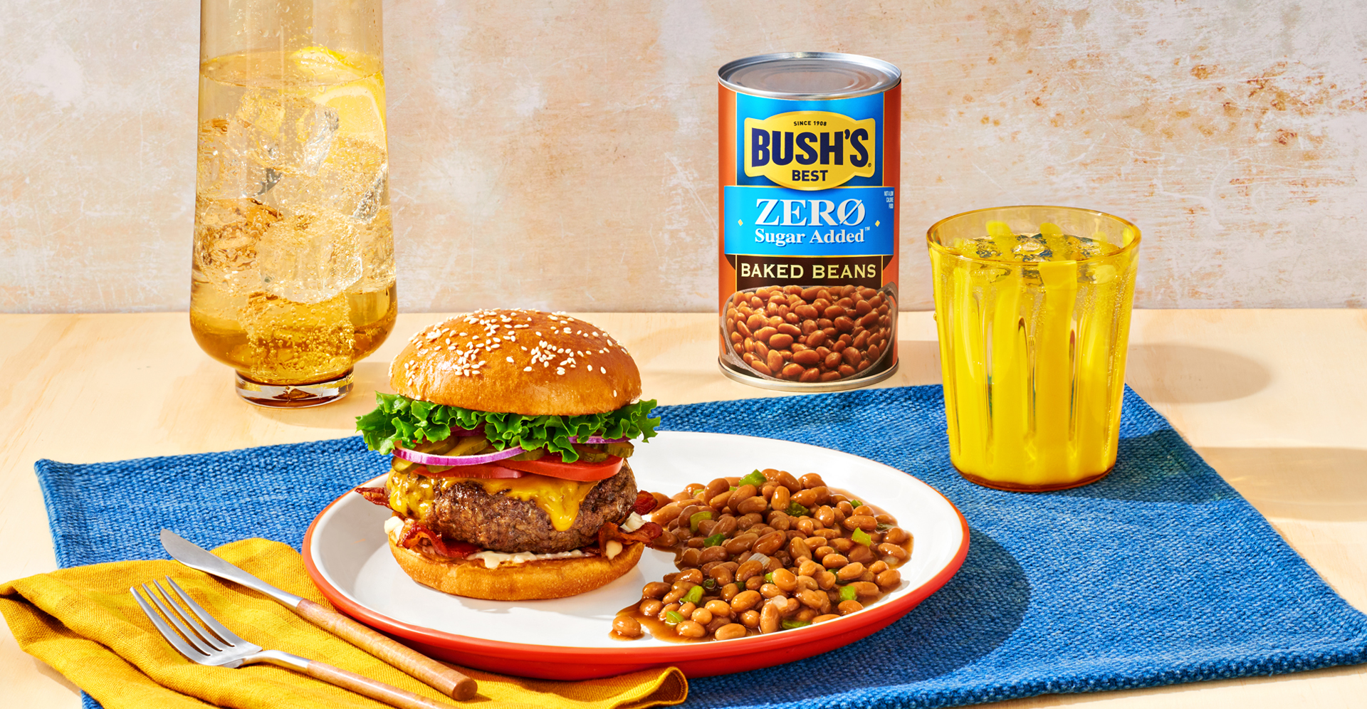 A plate with a burger and baked beans with a can of Bush's Zero Sugar Added Baked Beans on a picnic table.