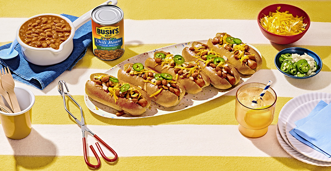 Long tray of chili bean dogs topped with jalapeños and cheddar, on a yellow and white striped tablecloth, surrounded by a can of Bush's chili beans and bowls of ingredients