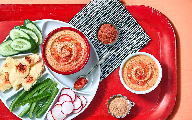 Tandoori Mango Hummus in bowl on red platter surrounded by spices, veggies, and naan.