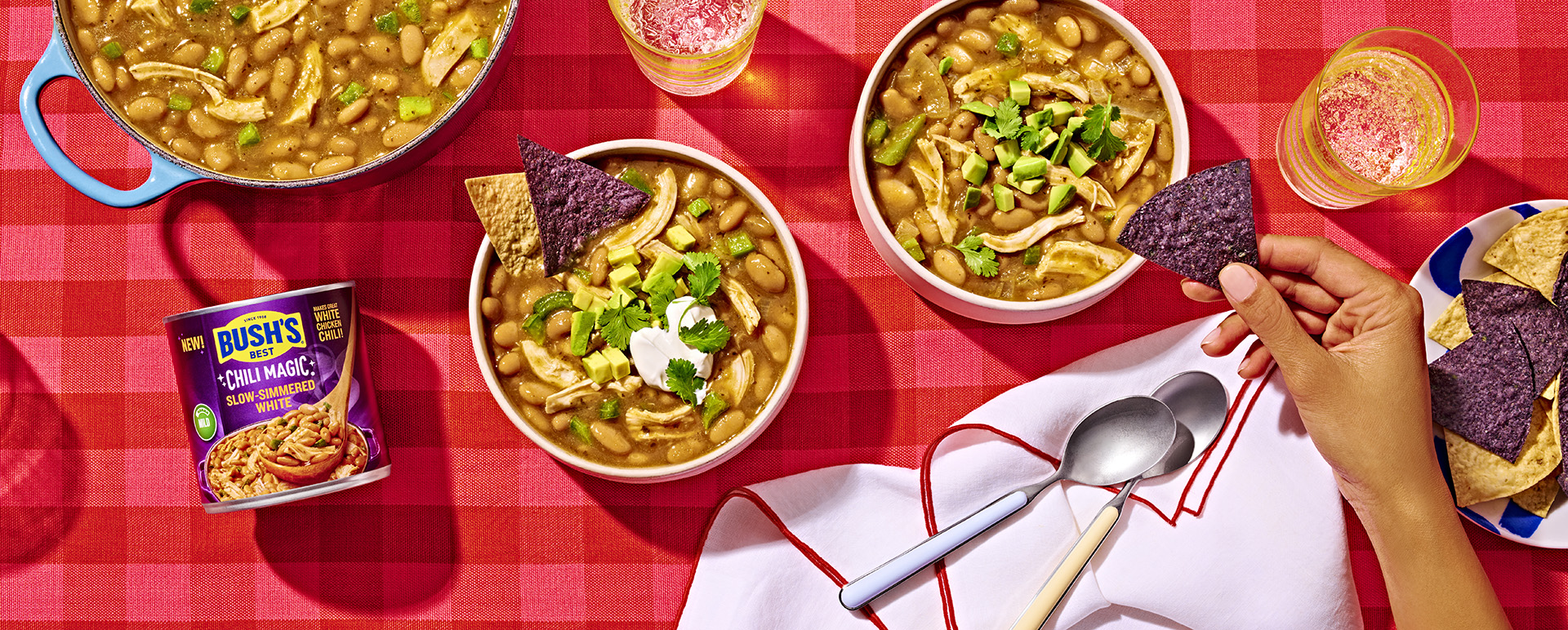 Two bowls of white chicken chili next to a can of Bush's Slow-Simmered White Chili Magic on a red tablecloth.