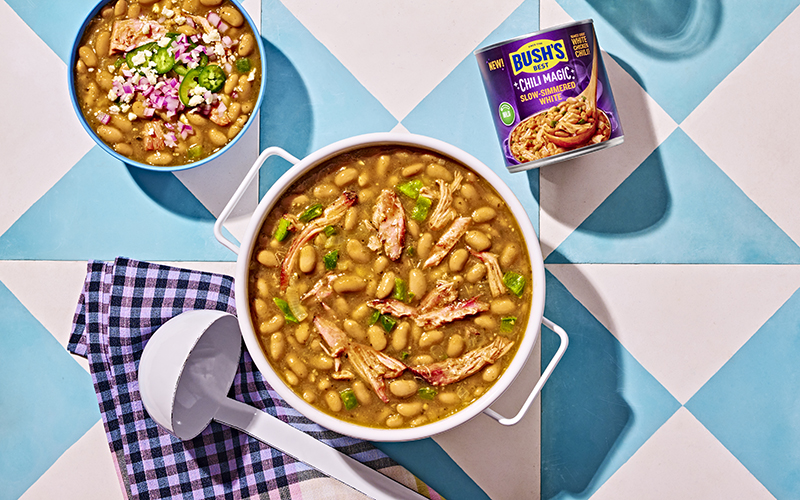 A bowl and a pot of chili verde with pork sitting next to a can of Bush's Slow-Simmered White Chili Magic