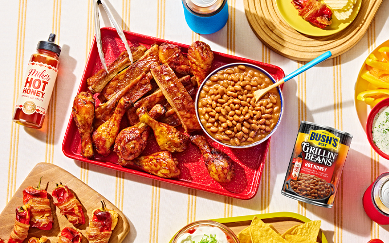 A bowl full of Bush's Hot Honey Grillin Beans on a red tray next to a pile of bbq chicken drumsticks and pork ribs.