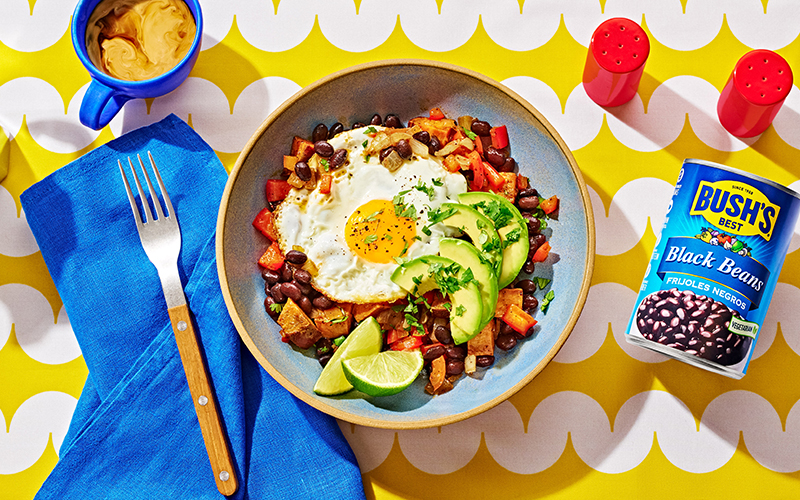 Bowl with colorful sweet potato and bean mixture, topped with a fried egg, avocado, and lime wedges
