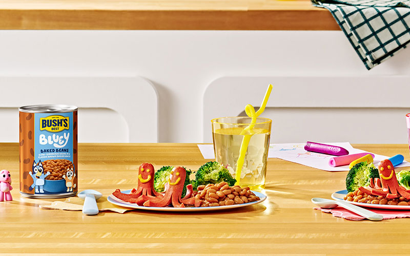 Plate with two octopus hot dogs with smiley faces drawn in mustard, Bush's Bluey baked beans, and broccoli.