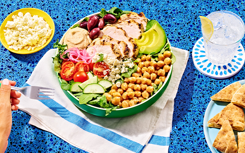 Mediterranean grain bowl salad in a green bowl on a white towel next to a plate with pita slices and a bowl of feta