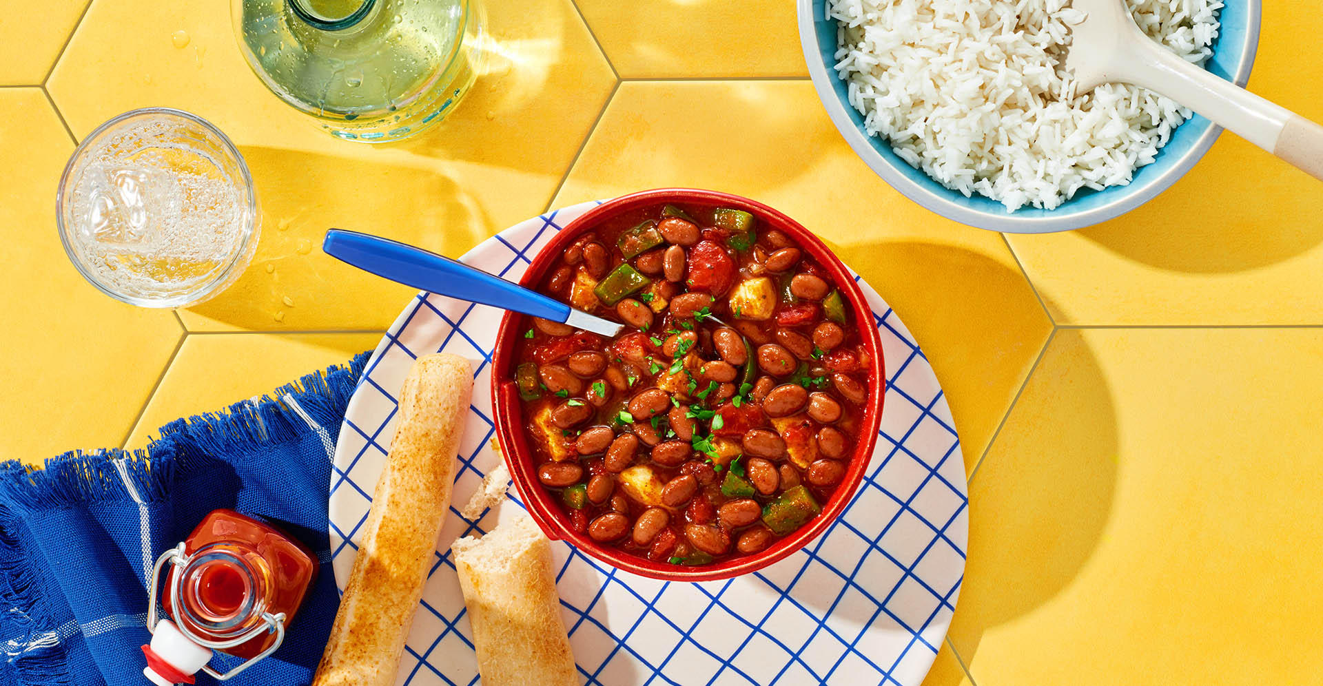 Red bowl of Hot Pepper Chicken Chili atop a yellow tile table with a side of rice and breadsticks.
