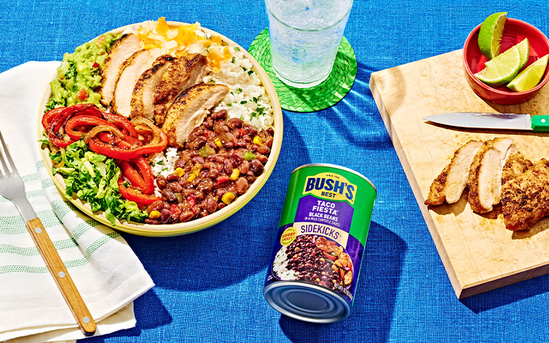 Yellow bowl full of lettuce, chicken, peppers, beans, rice, and cheese, next to a can of Taco Fiesta Sidekicks.