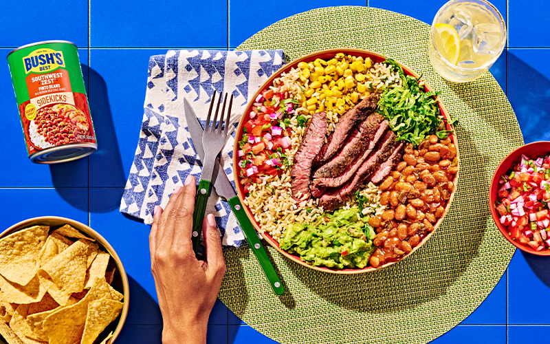 Steak burrito bowl surrounded by a can of Southwest Zest sidekicks, salsa, chips, jalapenos, and a drink