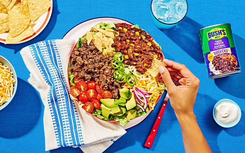 Red bowl full of lettuce topped with ground beef, Taco Fiesta beans, chips, cheese, tomatoes, avocado, jalapeños, and onions.