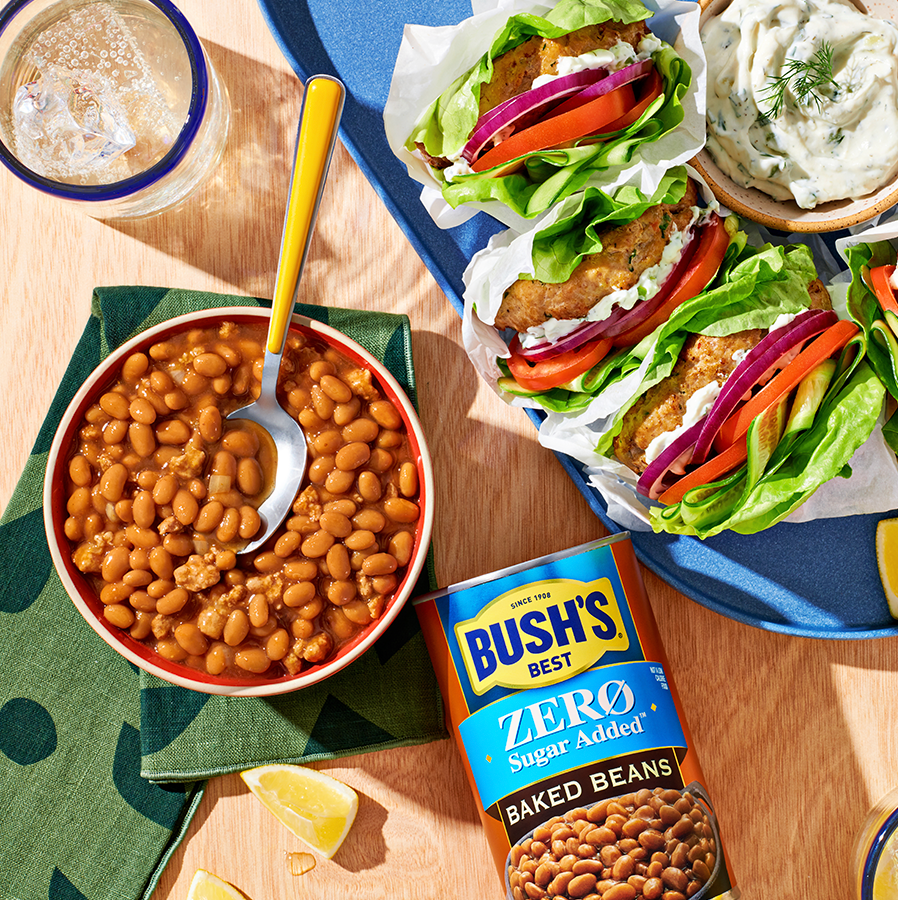 A bowl of baked beans on a set table next to a can of Bush's Zero Sugar Added baked beans and a plate of lettuce wraps.